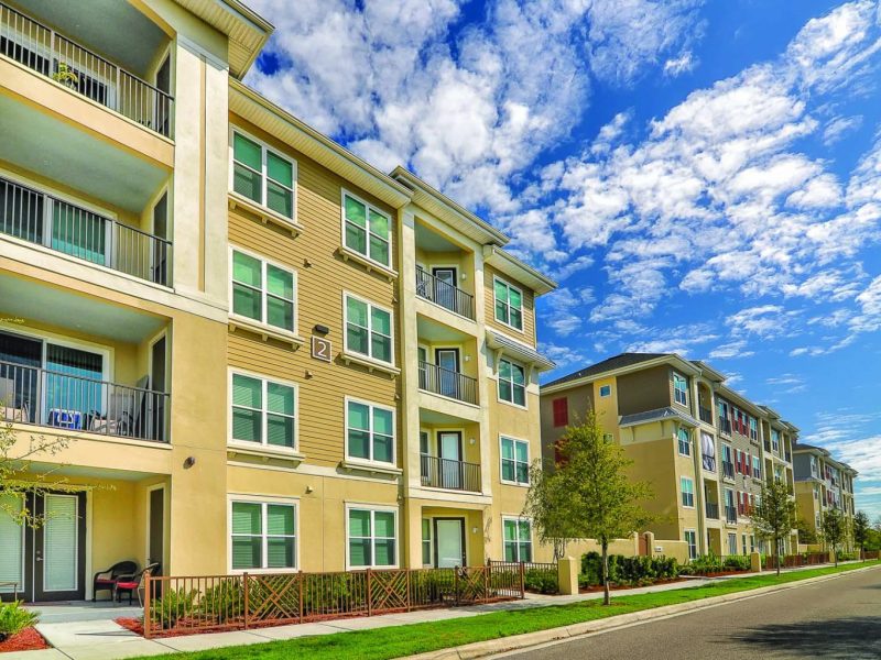 This image shows the TGM Ibis Walk Apartment aerial view, highlighting the exterior building's facilities concept. The trees, green grass, and the blue sky manifest the gorgeous environment.