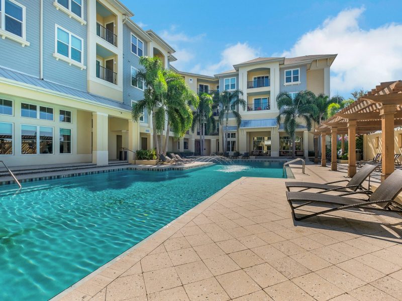 Modern apartment pool with palm trees, lounge chairs, and contemporary architecture under sunny sky.