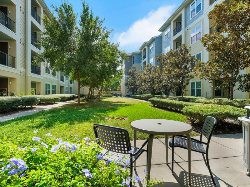 Beautiful residential courtyard with green landscaping, modern apartments, outdoor seating, and sunny blue sky.
